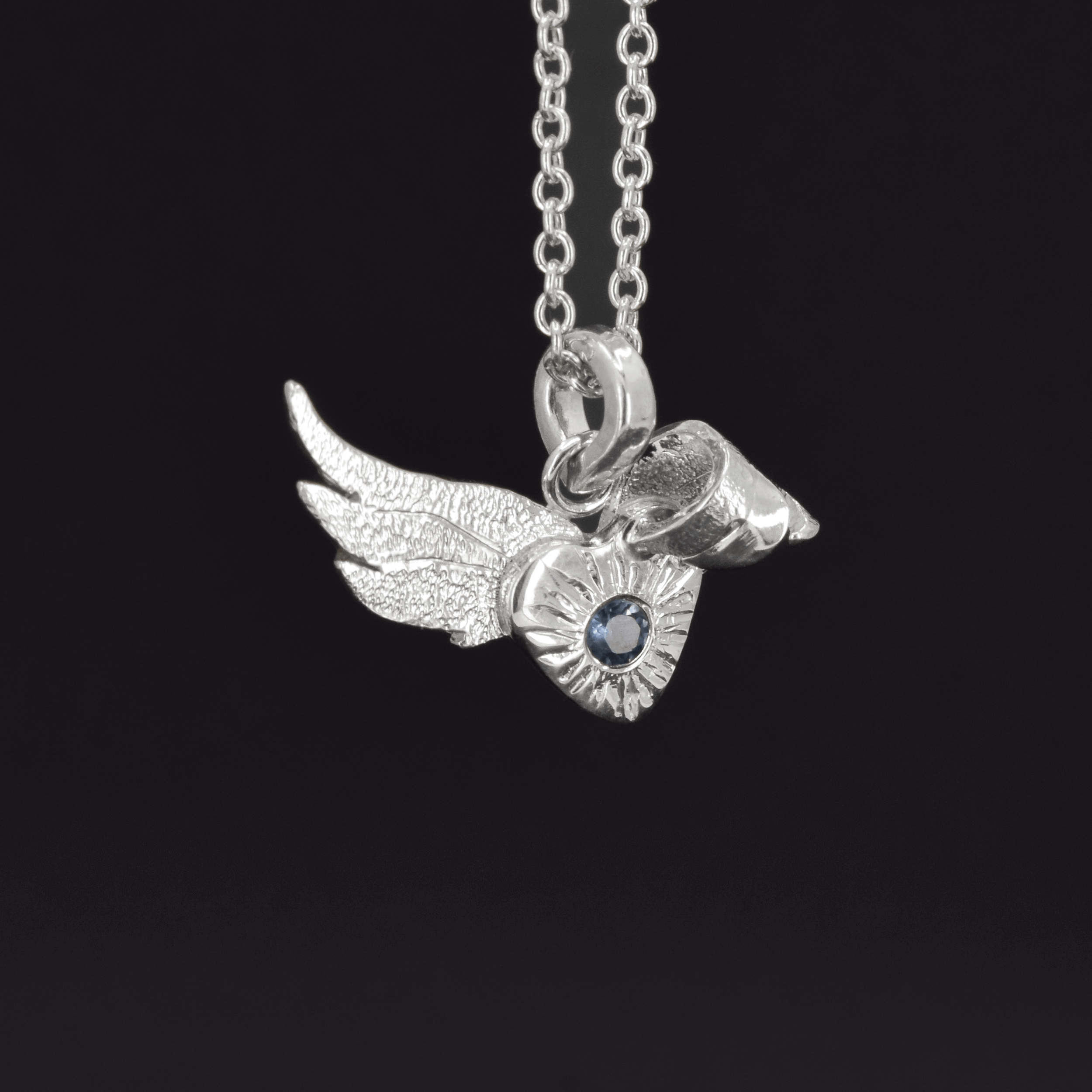 sterling silver angel wing pendant necklace with a flush set 4mm sapphire stone hanging from a cable chain. One of the wings is bent over the heart, which holds the stone.