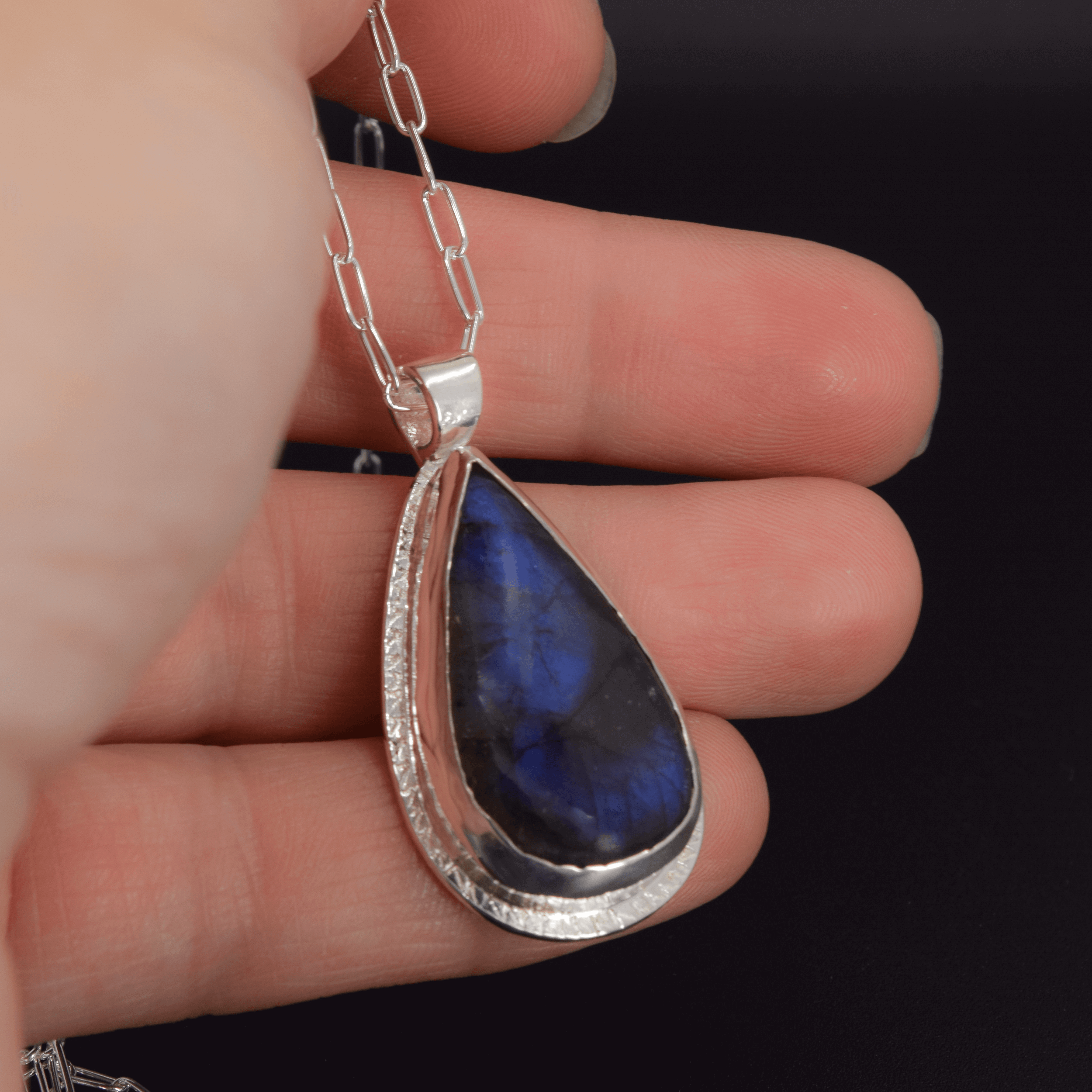 sterling silver pendant with a labradorite stone that has blue flashes hanging from a paperclip style chain held in hand for scale
