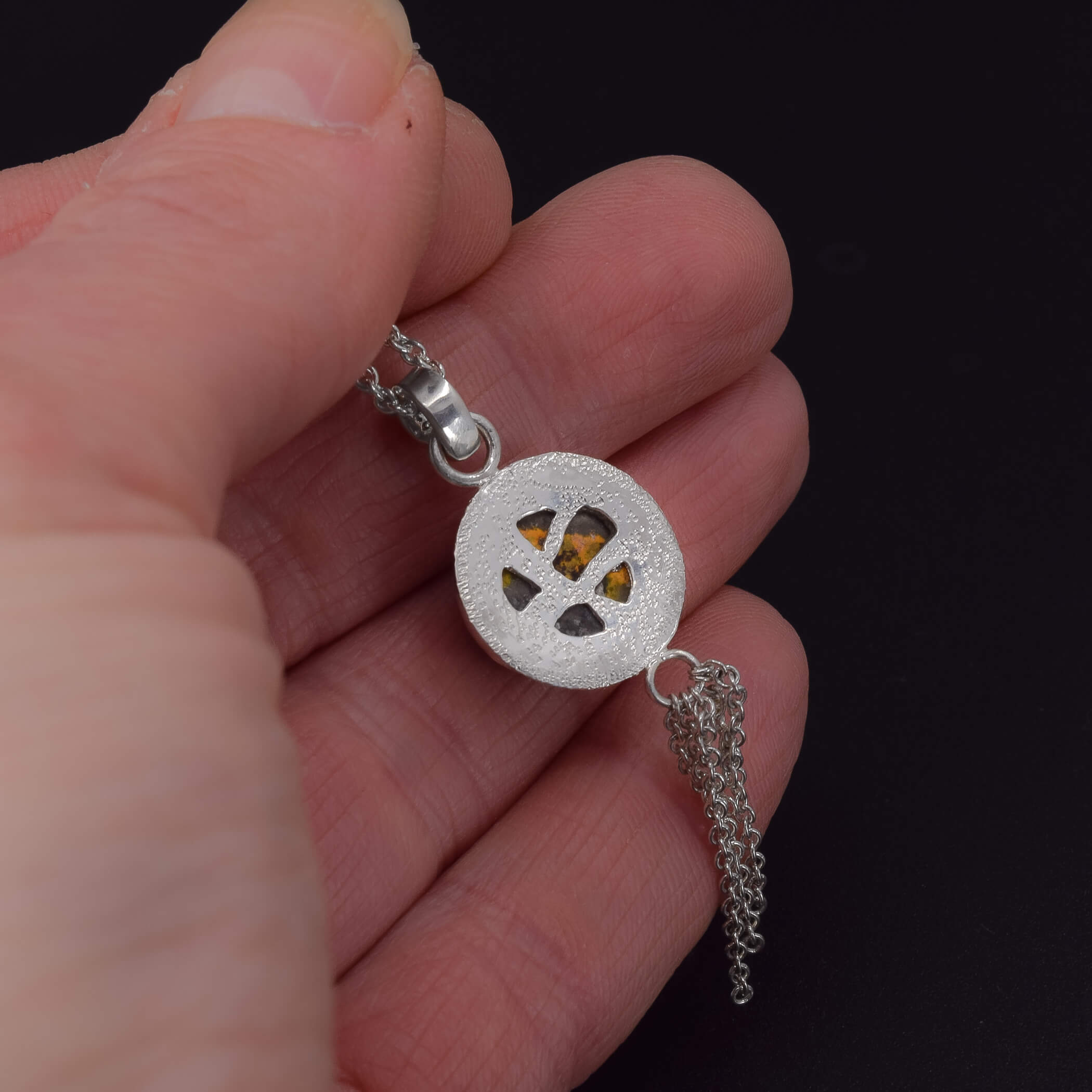 back of a sterling silver necklace featuring a round bumblebee jasper stone, with a pierced out design and a stardust texture shown in hand for scale