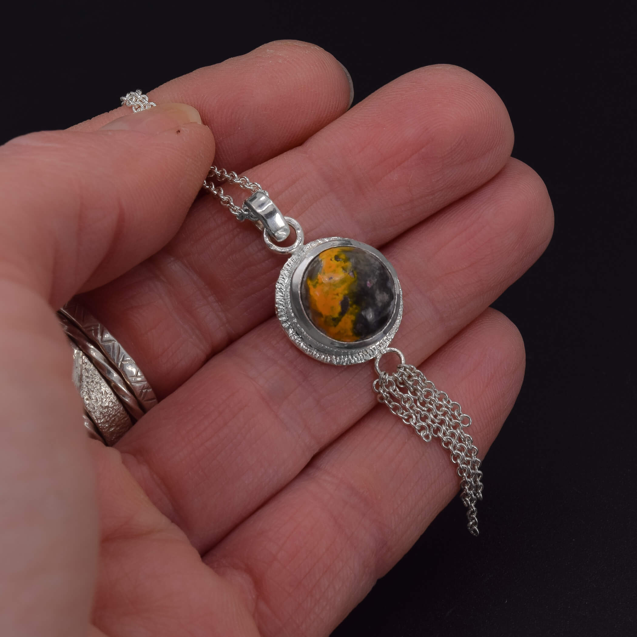 sterling silver necklace featuring a round bumblebee jasper stone, with a stardust texture around the edges and a chain embellishment at the bottom of the pendant shown in hand for scale