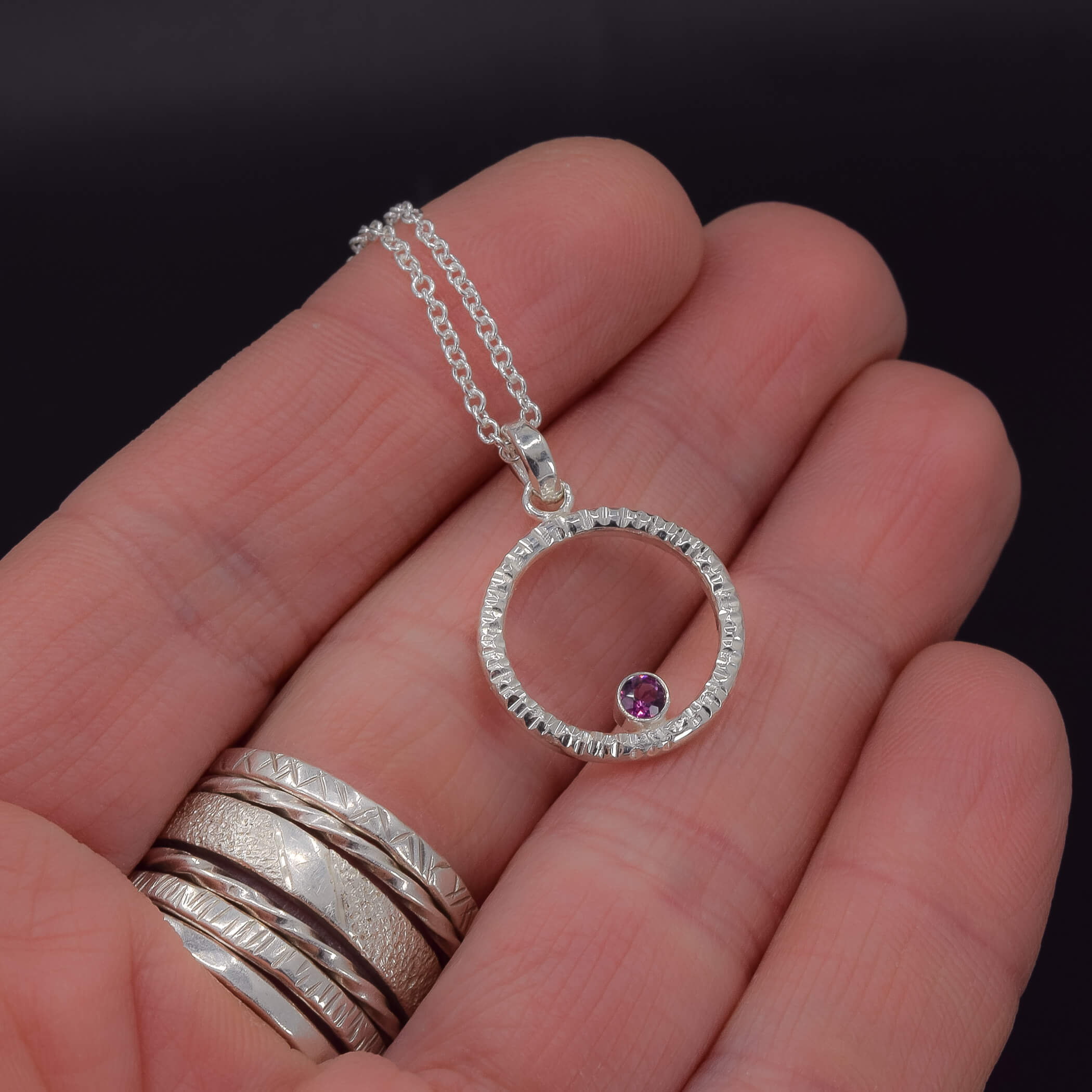Circle of life pendant necklace in sterling silver with textured edges, featuring a rhodolite garnet faceted stone shown in hand for scale