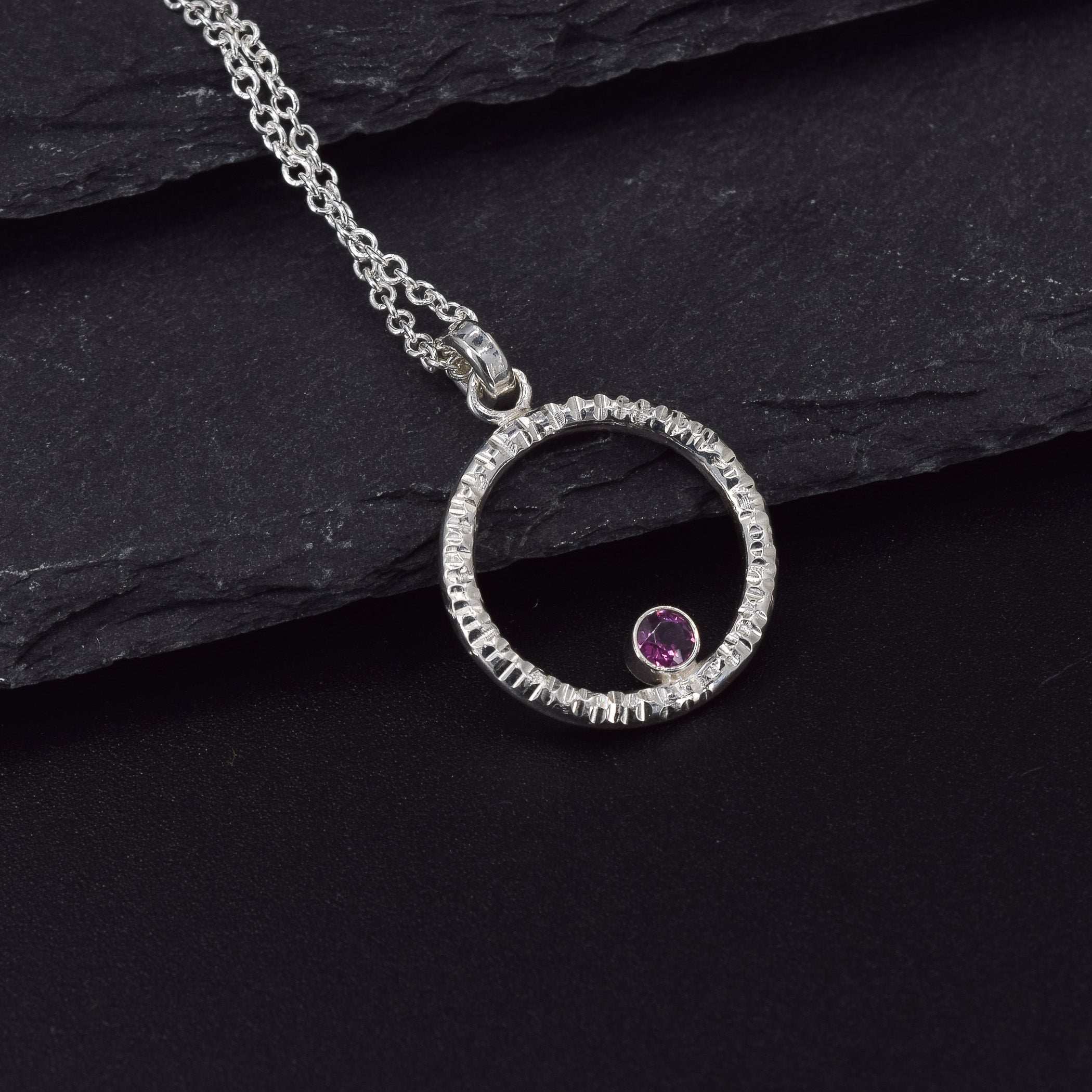Circle of life pendant necklace in sterling silver with textured edges, featuring a rhodolite garnet faceted stone leaning against a slate piece