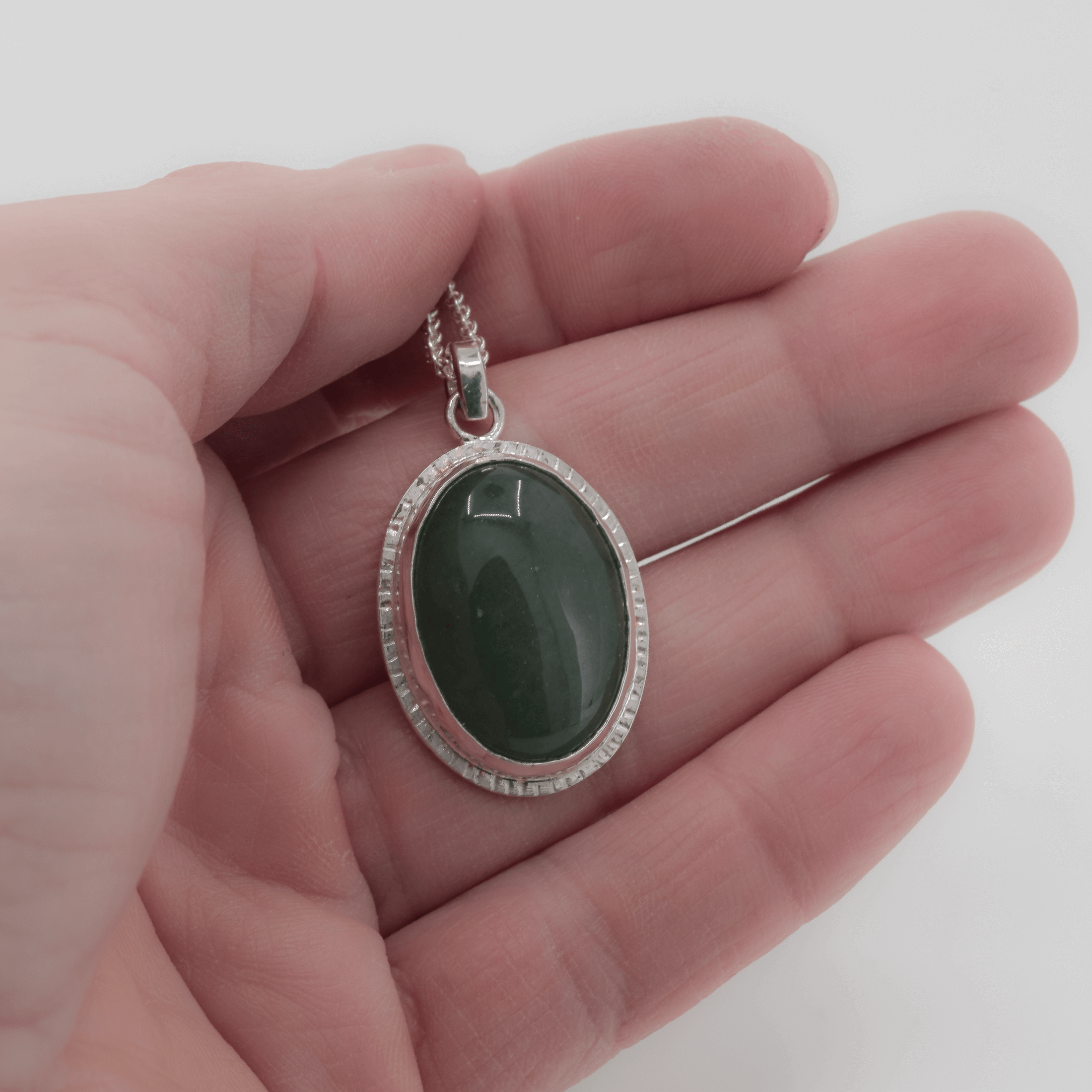 Green jade oval pendant with line textures around the edge of the bezel set stone shown in hand for scale