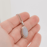 Oval moonstone pendant with a hand textured border hanging on a sterling silver cable chain shown in hand for scale