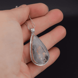 Teardrop shaped silver labradorite pendant with flashes of rainbow colors set in sterling silver, with a hand textured decorative border surrounding the stone and hanging from a paperclip style chain shown in hand for scale