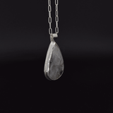Side view of a teardrop shaped silver labradorite pendant with flashes of rainbow colors set in sterling silver, with a hand textured decorative border surrounding the stone and hanging from a paperclip style chain