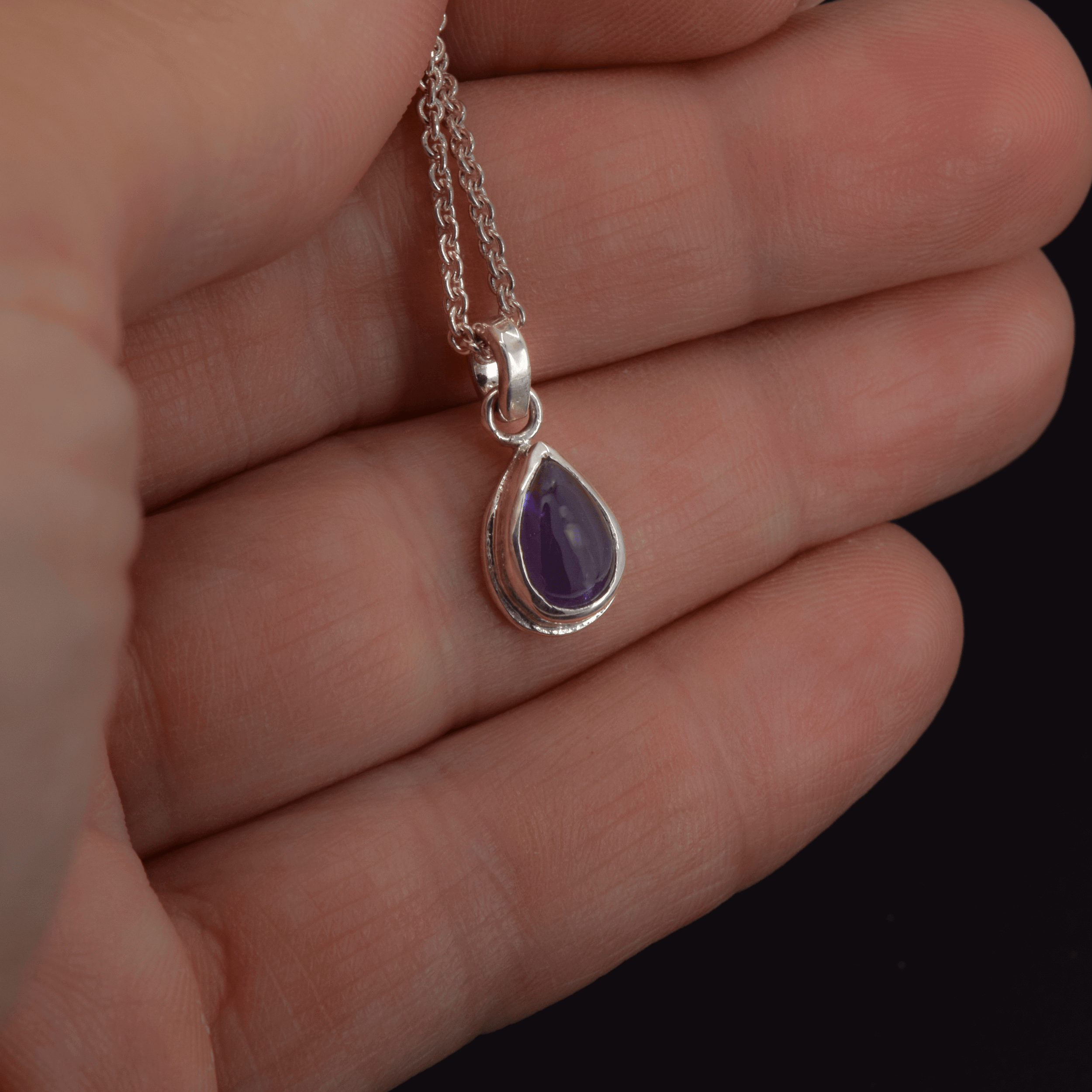 Teardrop shaped iolite pendant necklace in sterling silver hanging on a cable chain shown in hand for scale