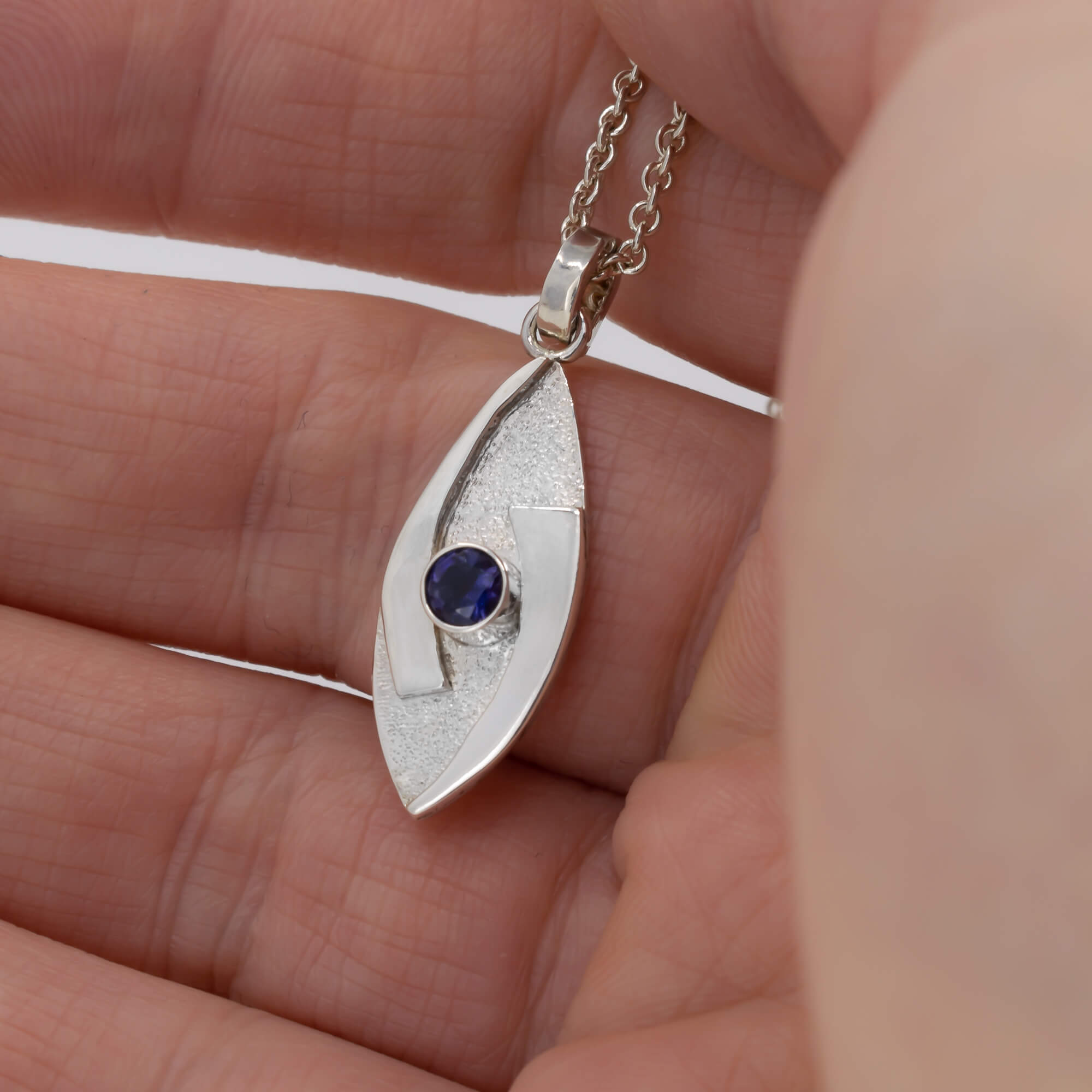 small hugged pendant necklace in sterling silver with a stardust texture and a 4mm Iolite  faceted stone shown in hand for scale