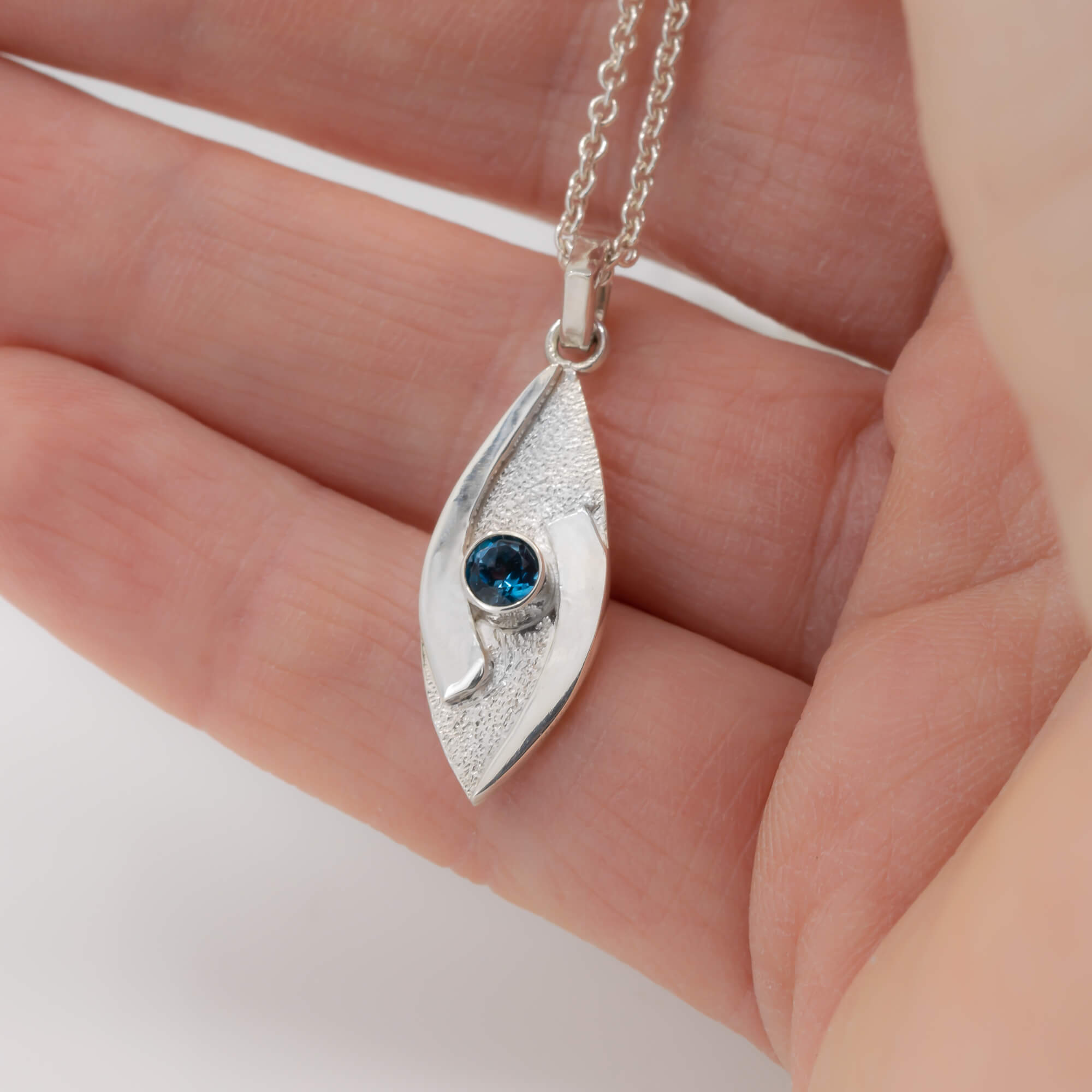 small hugged pendant necklace in sterling silver with a stardust texture and a 4mm London Blue Topaz faceted stone shown in hand for scale